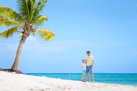 beautiful family of two, father and son, standing together at the beach with palm tree in the background enjoying summer vacation, white sand beach and turquoise lagoonの写真素材