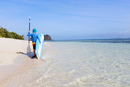 back view of little boy in rashguard holding surf board and paddle walking at the beautiful fiji island beach, active vacation concept, copy space on rightの写真素材