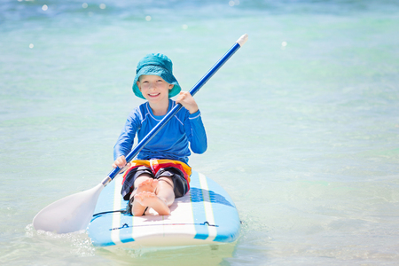 smiling little boy in rashguard holding paddle sitting at surf board enjoying tropical vacationの写真素材
