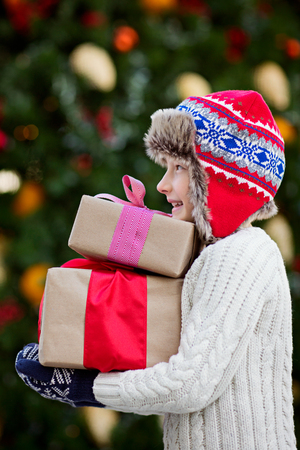 cute smiling little boy holding christmas presents with decorated tree in the background, magical christmas time conceptの写真素材
