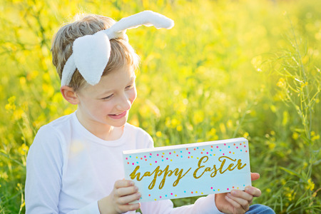 cute cheerful child wearing bunny ears and holding wooden sign happy Easter enjoying easter egg hunt...