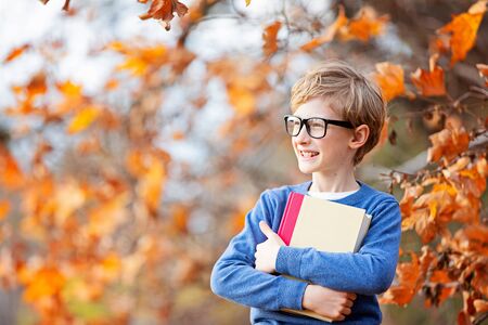 positive smiling boy in glasses holding book ready for a new school year, back to school concept, fall time in the park, copy space on leftの写真素材