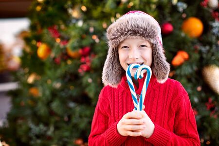 happy playful little boy holding candy canes by outdoor christmas tree, winter holiday conceptの写真素材