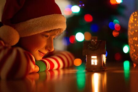happy little boy in santa's hat looking at candle light at cozy home and waiting for christmas miracle with tree lights and bokeh in the background, holiday concept, warm tonedの写真素材