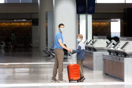 family of two, father and son, in face masks standing with the luggage in the airport, travel during coronavirus pandemic conceptの写真素材