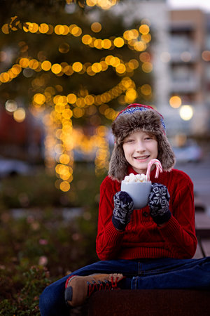 smiling boy holding cup with hot chocolate, marshmallows and candy canes enjoying outdoor holiday time with beautiful decorated with lights christmas tree in the background, vertical composition, family holiday conceptの写真素材