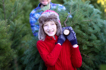 cheerful smiling boy and his dad carrying freshly cut tree at outdoor christmas tree farm, fun holiday family activity, copy space on leftの写真素材