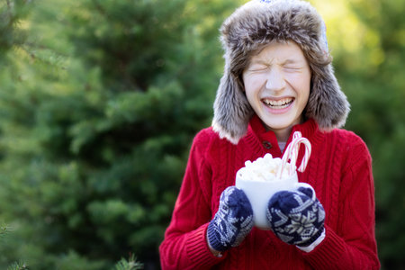 laughing cheerful boy holding cup with hot chocolate, marshmallows and candy canes and enjoying outdoor christmas time, copy space on left, holiday conceptの写真素材