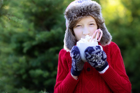 smiling cheerful boy holding cup with hot chocolate, marshmallows and candy canes and enjoying outdoor christmas time, copy space on left, holiday conceptの写真素材