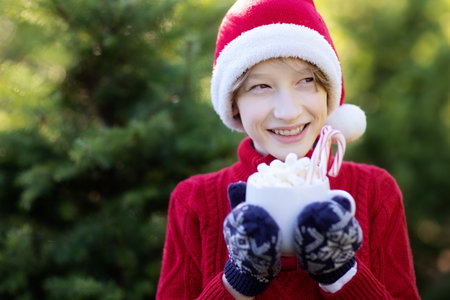 smiling cheerful boy in santa's hat holding cup with hot chocolate, marshmallows and candy canes and enjoying outdoor christmas time, copy space on left, holiday conceptの写真素材