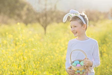 cute cheerful child wearing bunny ears and holding colorful basket full of easter eggs enjoying easter egg hunt at blooming flowers field, easter holiday concept, copy space on leftの写真素材