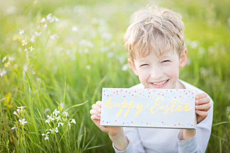 cute cheerful child holding wooden sign "happy Easter" enjoying easter egg hunt at blooming flowers field, easter holiday concept, copy space on leftの写真素材