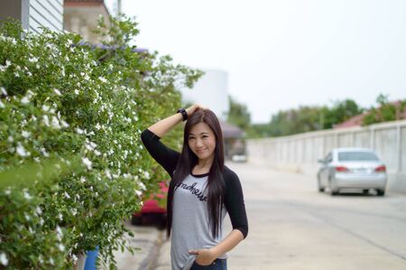 Portrait of lady attractive and smiling caucasian lady standing with Wearing a gray T-shirt Standing beside Bush Green. With white flowers on background of tree and wall blurry.の写真素材