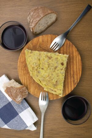 a piece of a Spanish omelet over a wood board with a fork some bread two glasses of wine and a piece of clothの写真素材