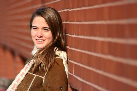 A teenage girl with braces leans against a red brick wall and smiles