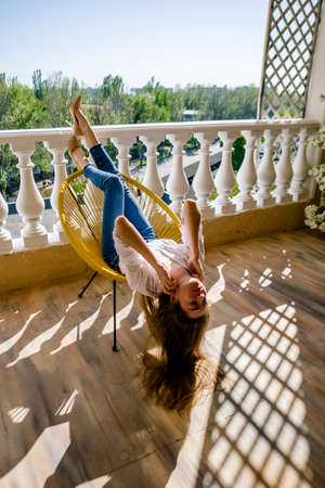 Woman relaxing and sunbathing at terrace. Young woman dressed in denim and white shirt, lying on chair, relaxing at home on balcony.の写真素材