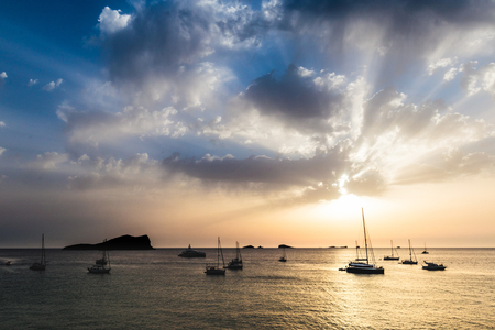 Rays of sun bursting through the sky clouds and over the sea during a sunset, casting strong colors and shadows over small islands and boatsの写真素材