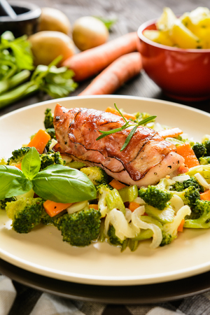 Baked chicken breast wrapped in bacon slices with boiled potatoes and steamed broccoli, carrot, garlic, onionの写真素材