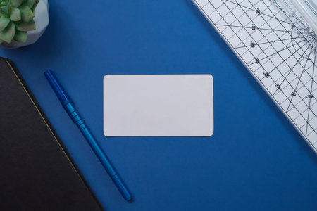 Overhead view of a blank white mockup card surrounded by blue pen, notebook, and ruler.