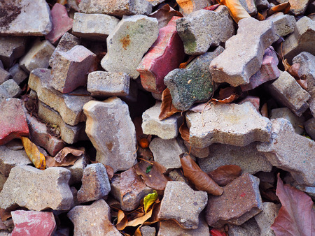 Randomly pile of broken cracked grainy texture red and grey worn out weathered worm jigsaw zigzag shape concrete block brick, dry yellow brown fallen leave backgroundの写真素材