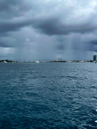 Heavy rain falls over a Florida marina with sailboats and yachts anchored offshore, dark storm...