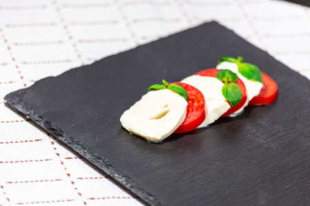 Caprese - mozzarella, tomato, green basil on a black slate board on a beautiful clothの写真素材