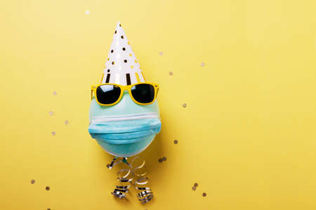 Concept of party during covid-19. Invitation card with blue ballon with protective mask, party hat and sunglasses. Top down view with copy spaceの写真素材