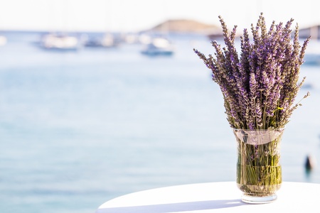 Lavender flowers in a glass vase on a table by the ocean.の写真素材