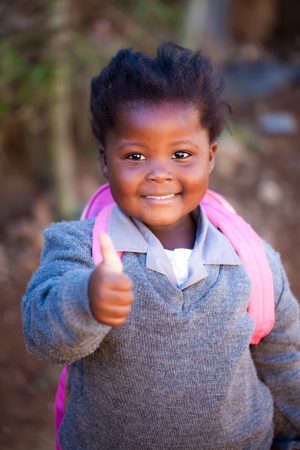 young african girl with a positiv thumb up on her way to school in a pink bag and gray school clothesの写真素材