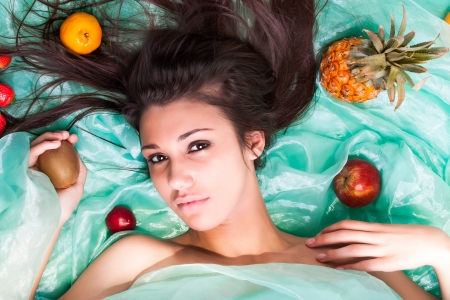 a close-up of a beautiful seductive young caucasion woman sprawled out in turqoise organza material surrounded by different colorful fruitの写真素材