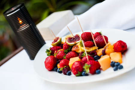 image of a porcelain plate filled with various fresh fruits and finger sticks on a white table and a burning candle in the backgroundの写真素材