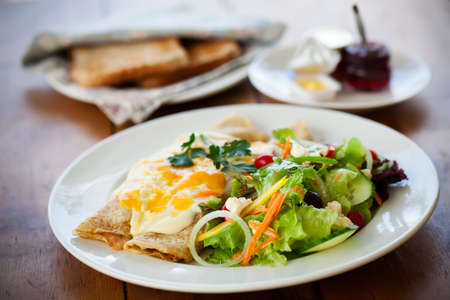 two filled crepes dribbled with cheese sauce plated with a healthy salad and some toast with condiments in the background all on a wooden tableの写真素材