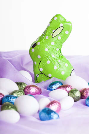 a green wrapped chocolate bunny placed on light purple organza material with various chocolate colored egss surrounding itの写真素材