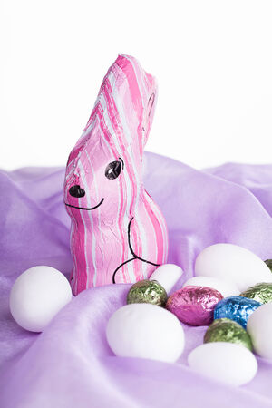 a chocolated moulded bunny wrapped in pink wrapping placed on purple organza material with various chocolate egss surrounding itの写真素材