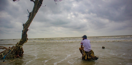 A young Man wearing white color t-shirt and a cap sitting on trunk of tree in front of sea beach in...