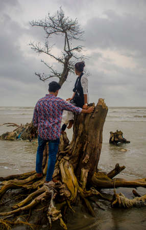 Girlfriend sitting on trunk of a broken tree and boyfriend standing by her at the Kuakata Sea beach in Patuakhali, Bangladesh before dusk.の写真素材