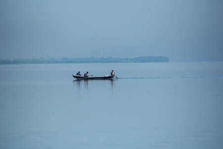 Three poor fishermen catching fish with fishing net from wooden row boat at blue water river of...