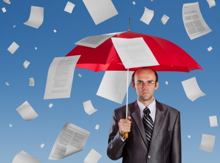 Serious businessman with red umbrella under falling documentsの写真素材