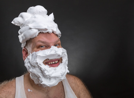 Bizarre smiling man with shaving foam on his face and on his head over grey backgroundの写真素材