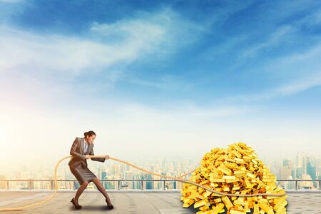 Businesswoman pulls the pile of gold bars against blue skyの写真素材