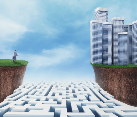 Businessman on the edge looks at the city situated on the other grass edge, there is a maze between two edgesの写真素材