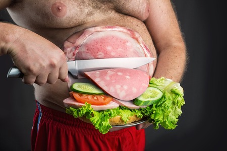 Fat man cutting a sausage slice for sandwich from his bellyの写真素材