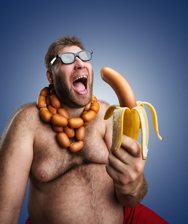 Mad man in glasses with sausages round his neck holds a big wurst in bananas peel over blueの写真素材