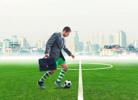 Businessman with suitcase in sportwear playing football in the town stadiumの写真素材
