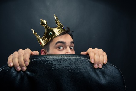 Scared man with crown on the head hidding behind the chairの写真素材