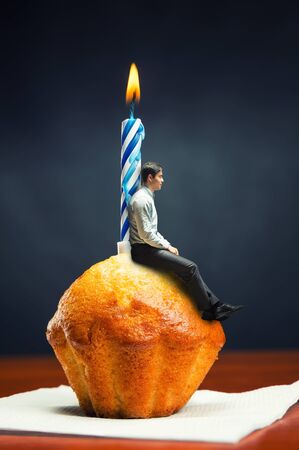 Sad birthday boy sits on cake with candle on the tableの写真素材