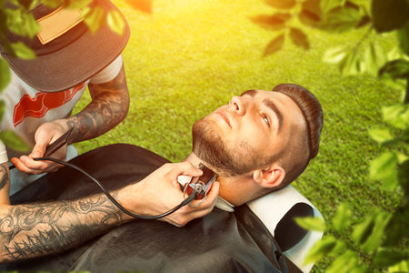 Young man having his beard shaven, barber working with trimmer over nature backgroundの写真素材