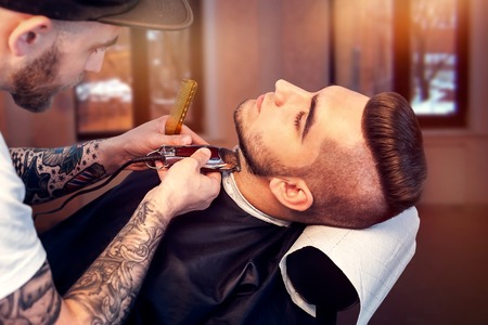 Young smiling man having his beard shaven, barber working with trimmerの写真素材