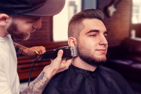 Young smiling man having his beard shaven, barber working with trimmer and combの写真素材