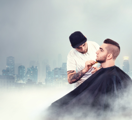 Young man having his beard shaven, barber working with comb over dark city backgroundの写真素材